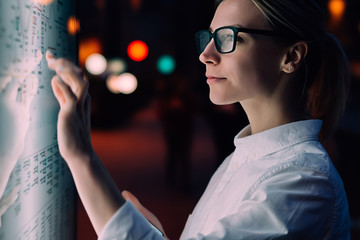 Interactive kiosk with public transport subway map.Female standing at big display with smartphone in hand.Young woman touching with finger screen while using train schedule application on mobile phone