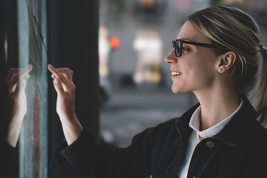 Smiling Female Standing At Big Display With Advanced Digital Technology. Young Woman Touching With Finger Sensitive Screen Of Interactive Kiosk For Find Information While Standing On Street In Evening