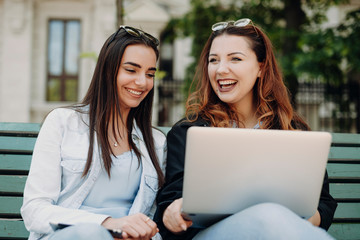Two caucasian beautiful female sitting on a bench laughing looking at a laptop against a building.