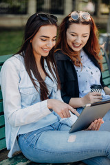 Side view portrait of a beautiful caucasian woman sitting on a beach and laughing looking to a tablet screen sitting with her girlfriend outside.