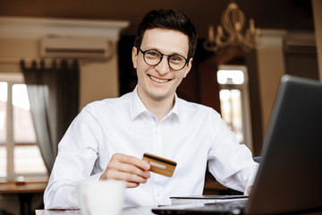 Portrait of a handsome young businessman looking at camera laughing while holding a gold credit card sitting at a desk working at his laptop.