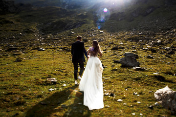 Back view of a unrecognizable bride and groom running in the mountains holding hands against sunrise.