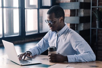 African business man using laptop, holding cup of coffee, sitting at his desk in modern loft office
