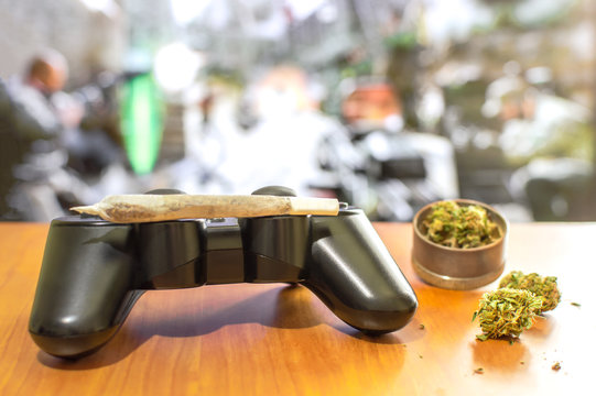 Video games and cannabis. Video game controller, big joint and grinder with marijuana buds on wooden table. Blur background of tv with video game.