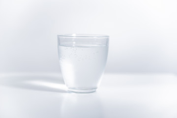 Round glass filled with cold mineral water on bright, blank surface. Reflection of the glass visible on the surface. High exposure photo.
