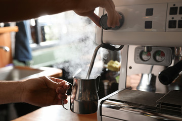 Barista steaming milk in metal jug with coffee machine wand at bar counter, closeup