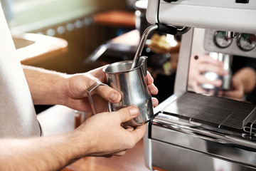 Barista frothing milk in metal pitcher with coffee machine wand at bar counter, closeup