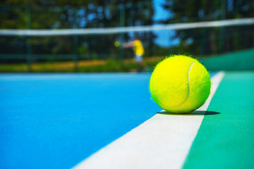Tennis ball on white court line on hard modern blue green court with player, net, balls, trees on the background. Close-up, selective focus. Sport, tennis play, healthy lifestyle concept.
