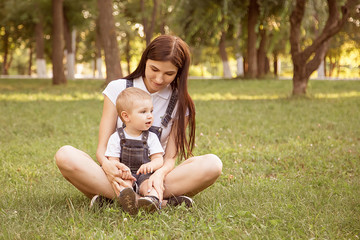 Fototapeta premium A young mother sits with her son, a boy in a lotus position in a park on a lawn, in the summer. Sunset sun, life style. A woman is dressed in a denim overall. Family look.
