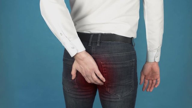 Man has pain due to Hemorrhoids. Concept of piles or hemorrhoidal disease in in the anal canal due to long-term work at the computer. Man in white shirt touches his buttocks with his hand. Red spot as