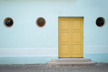 Baby blue wall with three small round windows and a yellow door without a handle. One way. Exit.