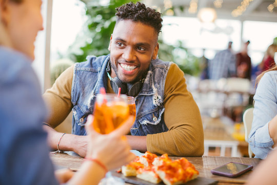Authentic image of happy African American guy eating pizza and drinking spritz cocktail with friends - multiracial couple or group of people dining and having fun in Italian restaurant