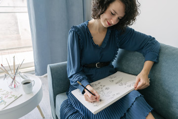 Elegant woman sitting on a sofa drawing flowers on a notebook at home