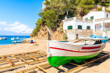 Traditional white and red colour fishing boat on Sa Riera beach with sea and village houses in background, Costa Brava, Spain