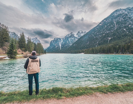 Back view of male with backpack standing on lake shore and fishing on sunny day in nature
