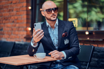 Attractive elegant businesman in sunglasses is sitting in cafe outside while chatting on mobile phone.