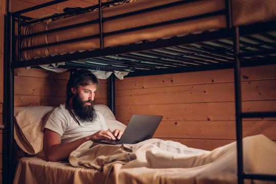 Concentrated bearded hipster working on laptop while lying in simple bunk bed of country wooden house