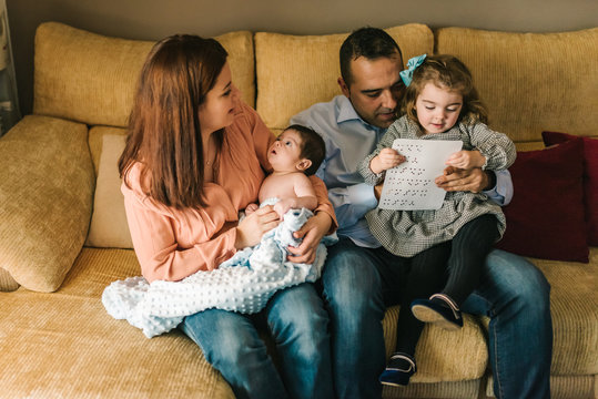 Happy young mother holding newborn baby wrapped in blanket and father holding daughter sitting on sofa at home