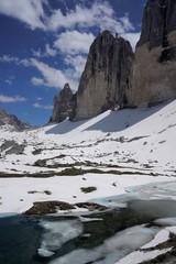 Obraz premium The peaks of Tre Cime di Lavaredo in Dolomites, Italy, blue sky with some clouds, lots of snow on the ground and partially frozen small lake in June. 