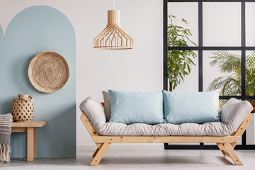 Wicker chandelier above wooden Scandinavian sofa with futon in bright living room interior