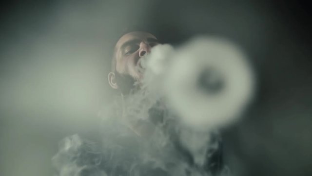 Close-up portrait of bearded brutal man vaping many small steam circles to camera on isolated black background in slow motion in professional dark studio. Round form of vapor is flying in air