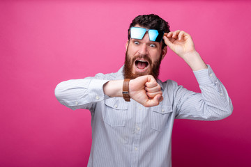 Young bearded man looking shocked at his watch, lifting up his sunglasses on pink background.