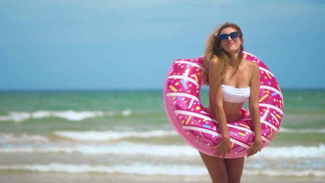 Woman having fun and smile dancing with pink donut. girl in bicini wearing sunglasses on the beach. Beautiful sexy woman on summer travel vacation.