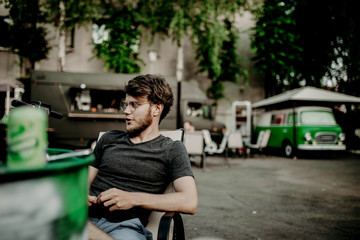 young hipster male with beard and glasses sits on street cafe on retro car background