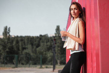 Close up portrait of young pretty caucasian fitness woman standing near the shipping container. Sportswoman drinking water outdoor. Breaking relax while exercise workout. Concept of health and sport.