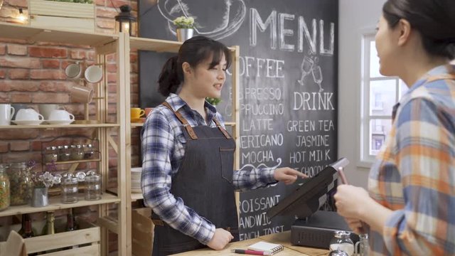 asian female regular customer patron making payment through mobile phone at counter in cafe bar. waitress barista in apron using pos machine checking order. bank app cellphone buying paying purchase