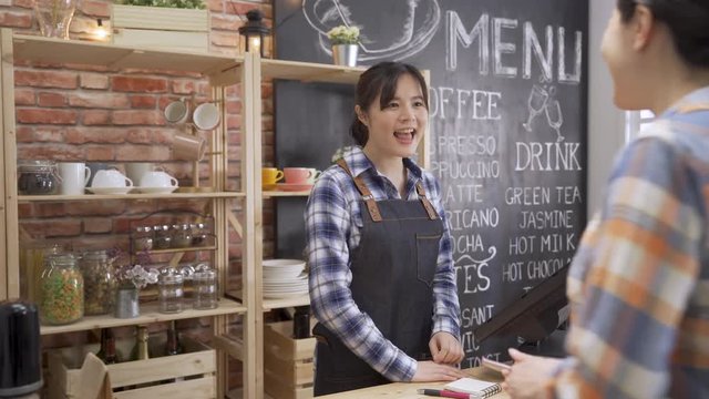 cheerful asian female bartender standing in counter in coffee shop welcoming regular customer in morning order on point of sale machine. service conversation in cafe bar. friendly waitress smiling.