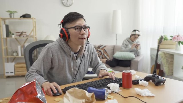 Noisy asian boyfriend plays video game on computer wear headset happy celebrating win raising hand clapping show share to girlfriend in back on sofa. lover indifferent ignore standoffish nod head