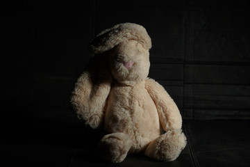 Soft toy rabbit on a dirty dark background in the beam of hard light with shadow. Lost childhood concept