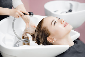 Concept beauty salon. Hairdresser washes hair of beautiful blonde girl under tap in wash