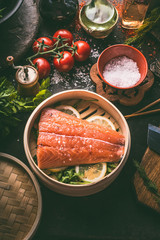 Raw salmon fillet in bamboo steamer on dark rustic kitchen table with fresh ingredients and tools....