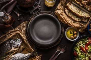 Food background of  Mediterranean food with Focaccia bread , roasted dorado fishes, salad bowl and olives oil on dark rustic kitchen table around empty plate. Delicious Mediterranean  cuisine concept.