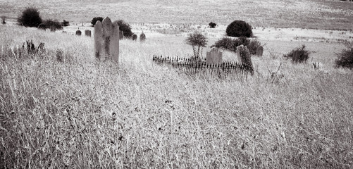 Tombstones in a graveyard overgrown by long grass sit forgotten on a lonely hillside. black and...
