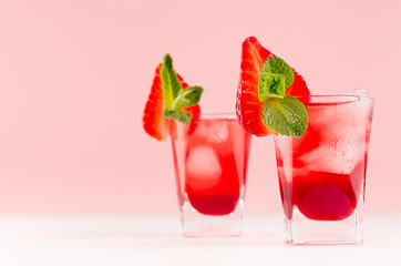 Summer fresh red bright drinks with ice cubes, strawberry slice, green mint on fashion pastel pink color background and white wood table, copy space.