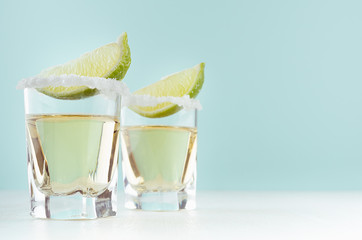Two tequila shots with salt rim and juicy piece lime on elegant white wood board and pastel mint...