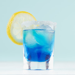 Cool summer blue beverage margarita for party with ice cubes, salt rim, lemon slice on white wood...