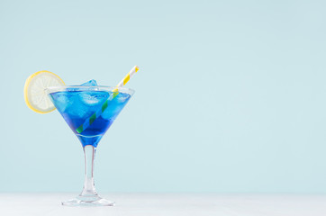 Cool summer blue beverage margarita for party with ice cubes,  lemon slice, yellow straw on white wood board and mint color wall.