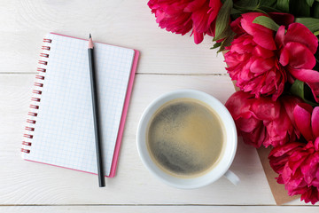Beautiful bright pink flowers peonies and a notebook with a cup of coffee on a white wooden background. top view.