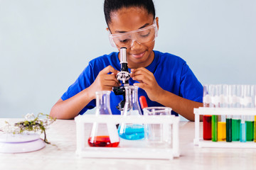 Joyful black beautiful cute clever girl in protective goggles sitting in school chemistry laboratory and looking into microscope