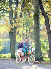 Fototapeta na wymiar Young active couple, bearded man and attractive long-haired blond woman riding tandem bicycle along asphalt path in lit by bright sun beautiful park under tall trees with green and yellow leaves.