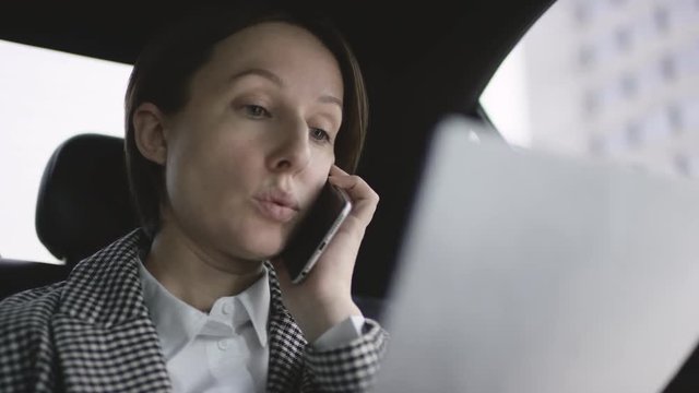 woman with brown hair in white blouse and check coat riding in car and discuss work documents by her phone