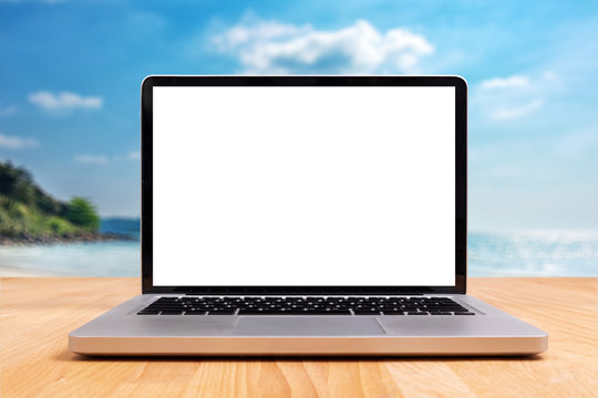 laptop computer with empty blank white screen for copy space on wooden desk with blurry summer sea ocean beach at background