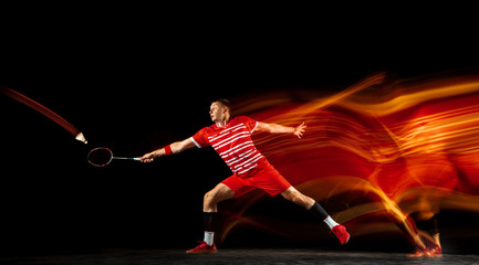 Young man playing badminton isolated on black background in mixed light. Male model with the racket in action, motion in game with the fire shadows. Concept of sport, movement, healthy lifestyle. - obrazy, fototapety, plakaty
