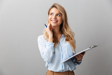 Photo of alluring blond secretary woman with long curly hair thinking and holding clipboard while working in office - obrazy, fototapety, plakaty