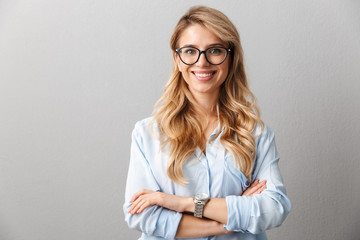 Photo of happy blond businesswoman wearing eyeglasses smiling and standing with hands crossed - obrazy, fototapety, plakaty