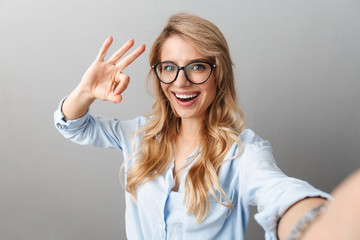 Photo of pretty blond businesswoman wearing eyeglasses smiling and showing ok sign while taking...